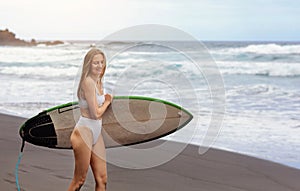 Young surfer woman in bikini going surfing stands with surfboard on the black sandy beach. Girl walks with longboard