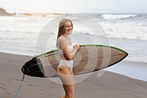 Young surfer woman in bikini going surfing stands with surfboard on the black sandy beach. Girl walks with longboard