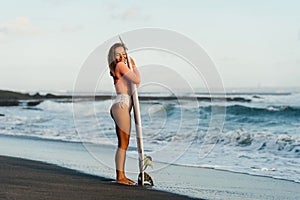 Young surfer woman holding white short surfboard on a beach at sunset or sunrise.