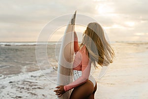 Young surfer woman holding white short surfboard on a beach at sunset or sunrise.