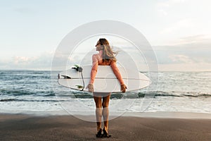 Young surfer woman holding white short surfboard on a beach at sunset or sunrise.