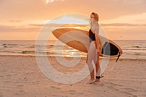 Young surfer woman with surfboard on beach and warm sunrise
