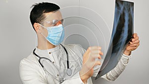 A young surgeon wearing a protective medical mask examines an X-ray of a patient's lungs and ponders the diagnosis. A