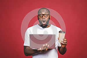 Young surprised african man standing and using laptop computer isolated over red background. Drinking coffee or tea