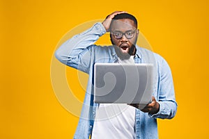 Young surprised african man standing and using laptop computer isolated over yellow background