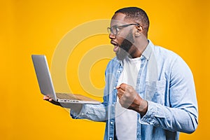 Young surprised african man standing and using laptop computer isolated over yellow background