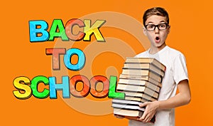 Young surprised boy in glasses holding many books on orange