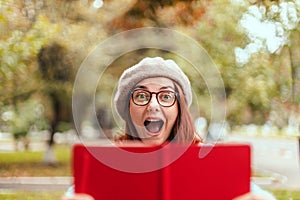 Young surprised girl in a beret, eyeglasses and a warm sweater holds a red notebook in the autumn park