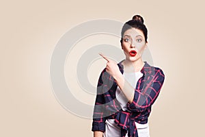 Young surprised girl with casual style and bun hair pointing her finger sideways, demonstrating something on beige blank wall