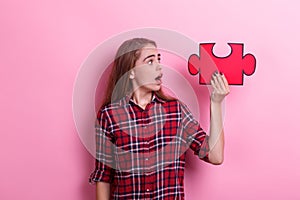 A young surprised girl holds a large puzzle on raised hand and looks at it. On a pink background.