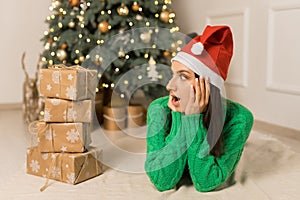 The young surprised girl lying near Christmas festive tree and brown gifts