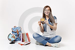 Young surprised joyful woman student in glasses denim clothes holding credit card sitting near globe backpack, school