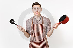 Young surprised man chef or waiter in striped brown apron, shirt holding red empty stewpan black ladle isolated on white