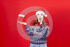 young surprised woman with gift Shoe in her hand in a sweater with snowflakes and a Santa Claus hat on a red background