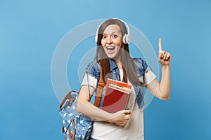 Young surprised woman student with backpack headphones listening music hold school book pointing index finger up on copy