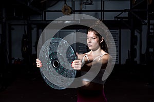 Young sweaty muscular strong fit girl holding big heavy barbell weight plate with her hands and lifting it up as hardcore cross wo