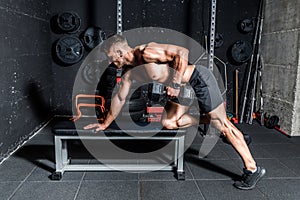 Young sweaty muscular strong man doing workout training back muscles with dumbbell on the bench in the gym dark image real people