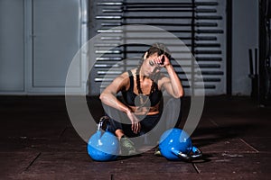 Young sweaty strong muscular fit girl with big muscles sitting on the floor and with two heavy kattlebell weights taking a break r