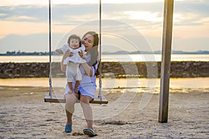 Young sweet and happy Asian Chinese woman holding baby girl swinging together at beach swing on Summer sunset in mother and little