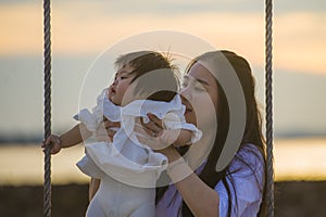 Young sweet and happy Asian Chinese woman holding baby girl swinging together at beach swing on Summer sunset in mother and little