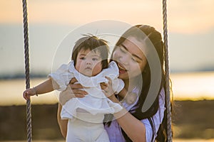 Young sweet and happy Asian Chinese woman holding baby girl swinging together at beach swing on Summer sunset in mother and little