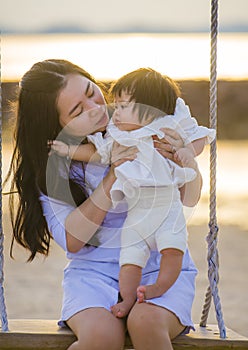 Young sweet and happy Asian Chinese woman holding baby girl swinging together at beach swing on Summer sunset in mother and little