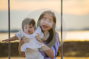 Young sweet and happy Asian Chinese woman holding baby girl swinging together at beach swing on Summer sunset in mother and little