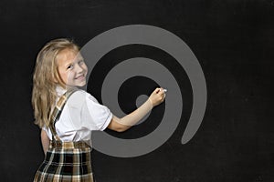 Young sweet junior schoolgirl with blonde hair smiling happy writing with chalk in blackboard