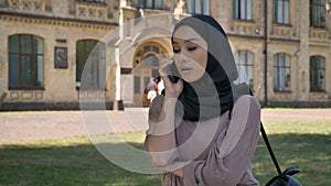 Young sweet muslim girl in hijab is talking on phone and hand up in daytime in summer, building on background, religiuos