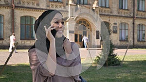 Young sweet muslim girl in hijab is talking on phone and laughing in daytime in summer, building on background