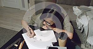 Young talented female artist drawing pencil sketches sitting on the floor in the evening at home