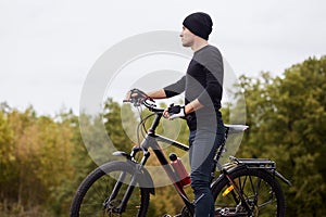 Young tall athletic man standing with bicycle in autumn park, looking far away, holding handlebars, wearing special clothes,