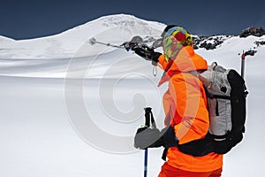 A young tall professional skier points with a ski stick to the top of the mountain while standing high in the mountains.