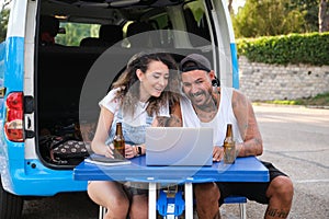 Young tattooed couple smile and talk in a laptop video call during a van trip.