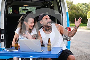 Young tattooed couple smile and talk in a laptop video call during a van trip.