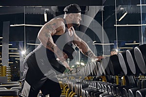 Young, tattooed, muscular man in black shorts, vest and cap. Exercising with dumbbells for training his triceps in dark