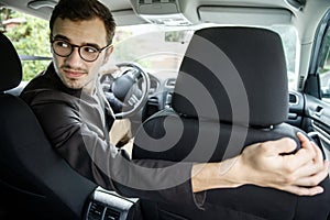 Young taxi driver looks back over the seats. His right hand is on the seat. He is sitting at his car