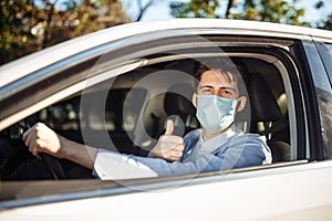 Young taxi driver shows thumb up sign sitting in the car and wearing protective sterile medical mask, works hard during