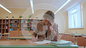 A young teacher female sits alone at a desk in a school class and checks a stack of students' notebooks