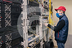 Young technician in medical mask switches fiber optic wires on the main router. The installer works in the server room during