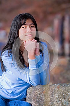 Young teen biracial girl quietly resting elbows on rock along lake shore