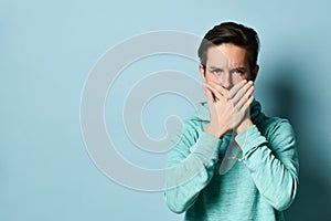 Young teen boy in blue hoodie standing and closing mouth to keep silence over pastel blue wall background