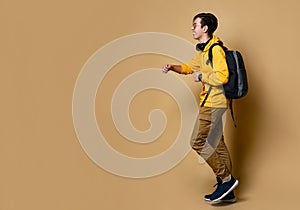 Young teen boy in comfortable clothing, sneakers and backpack jumping over yellow wall background