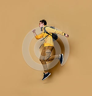Young teen boy in comfortable clothing, sneakers and backpack jumping over yellow wall background