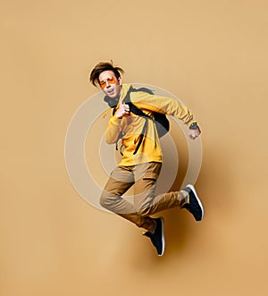 Young teen boy in comfortable clothing, sneakers and backpack jumping over yellow wall background