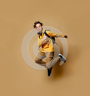 Young teen boy in comfortable clothing, sneakers, sunglasses and backpack jumping and feeling cool over yellow wall background