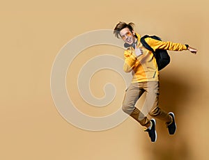 Young teen boy in comfortable clothing, sneakers, sunglasses and backpack jumping and feeling cool over yellow wall background