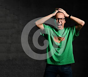 Young teen boy in green t-shirt, jeans and glasses standing, holding head, screaming and feeling shock over dark background