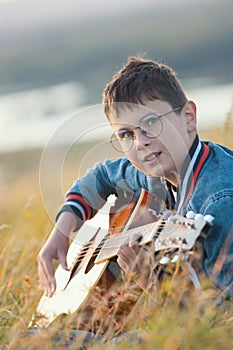 Young teen boy playing on acoustic guitar at summer field on sunset and looking to camera
