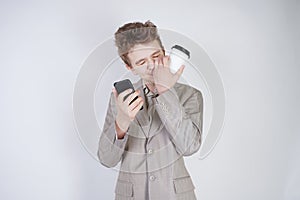 Young teen caucasian student standing in grey business suit, calling by smart phone, drinking coffee, touching his nose and lying